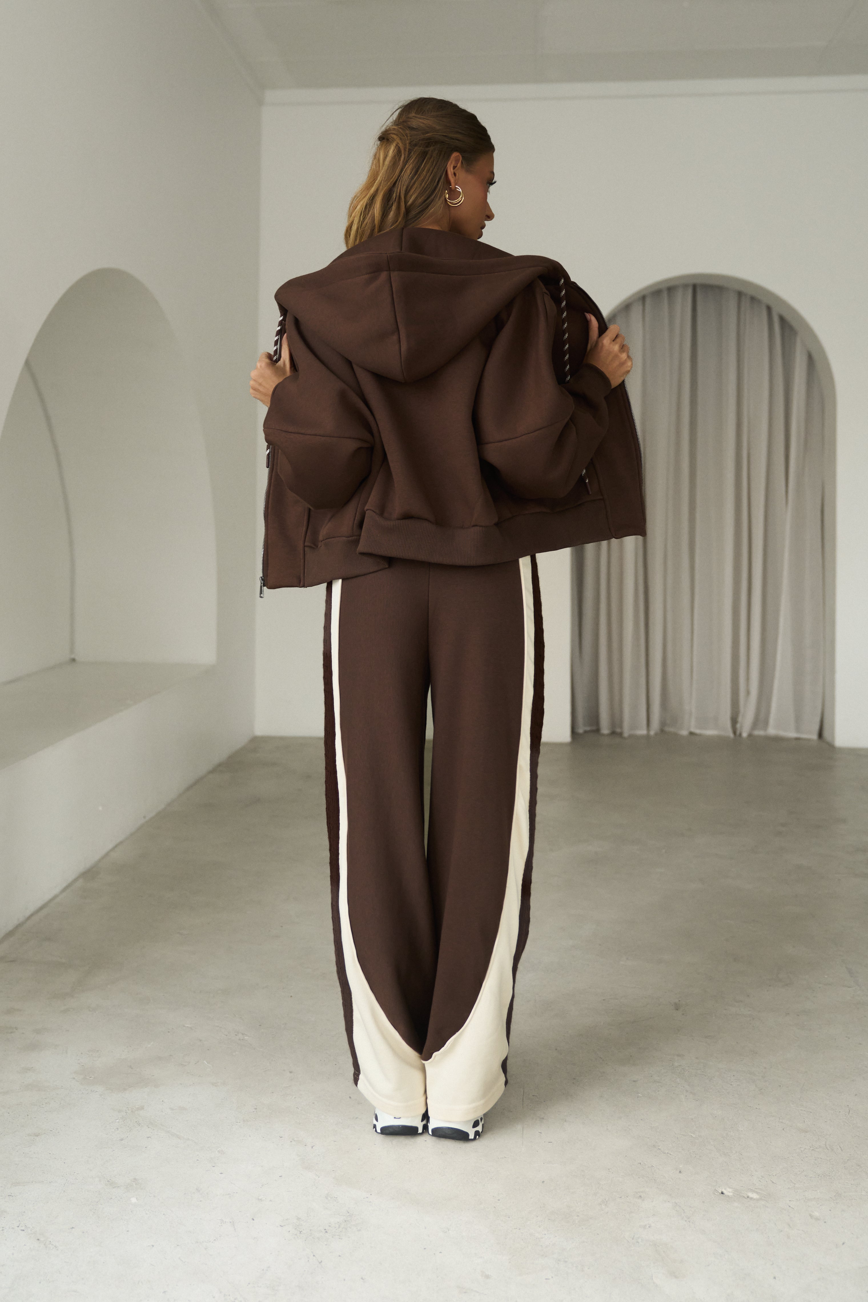 A model viewed from behind wearing a dark brown cropped hoodie and matching wide-leg sweatpants with cream side stripes, standing in a minimalist white room with concrete floors.