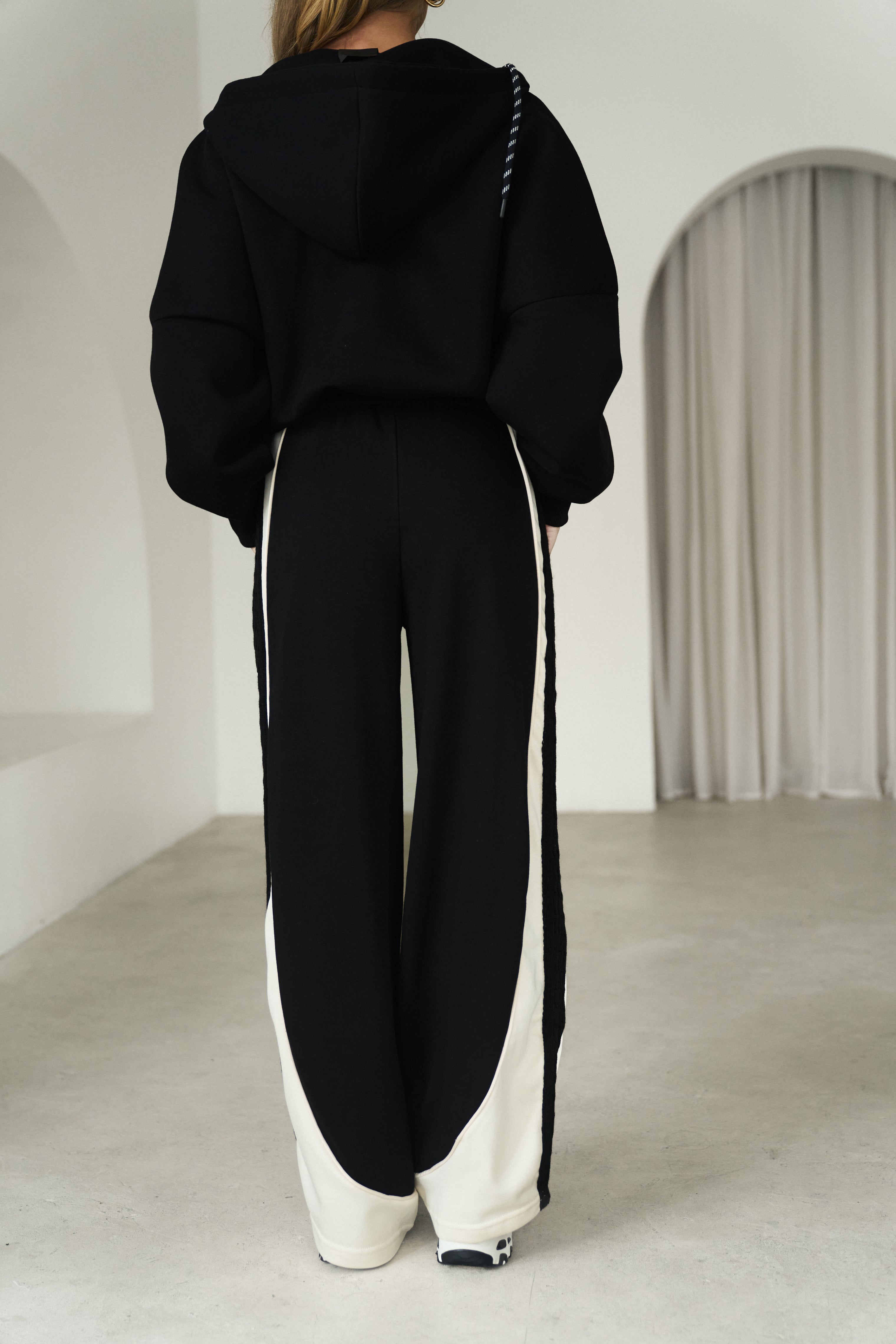 A person shown from the back wearing a black cropped hoodie and matching wide-leg pants with bold off-white side panels and stripes, standing in a bright minimalist interior with white walls and a draped curtain.