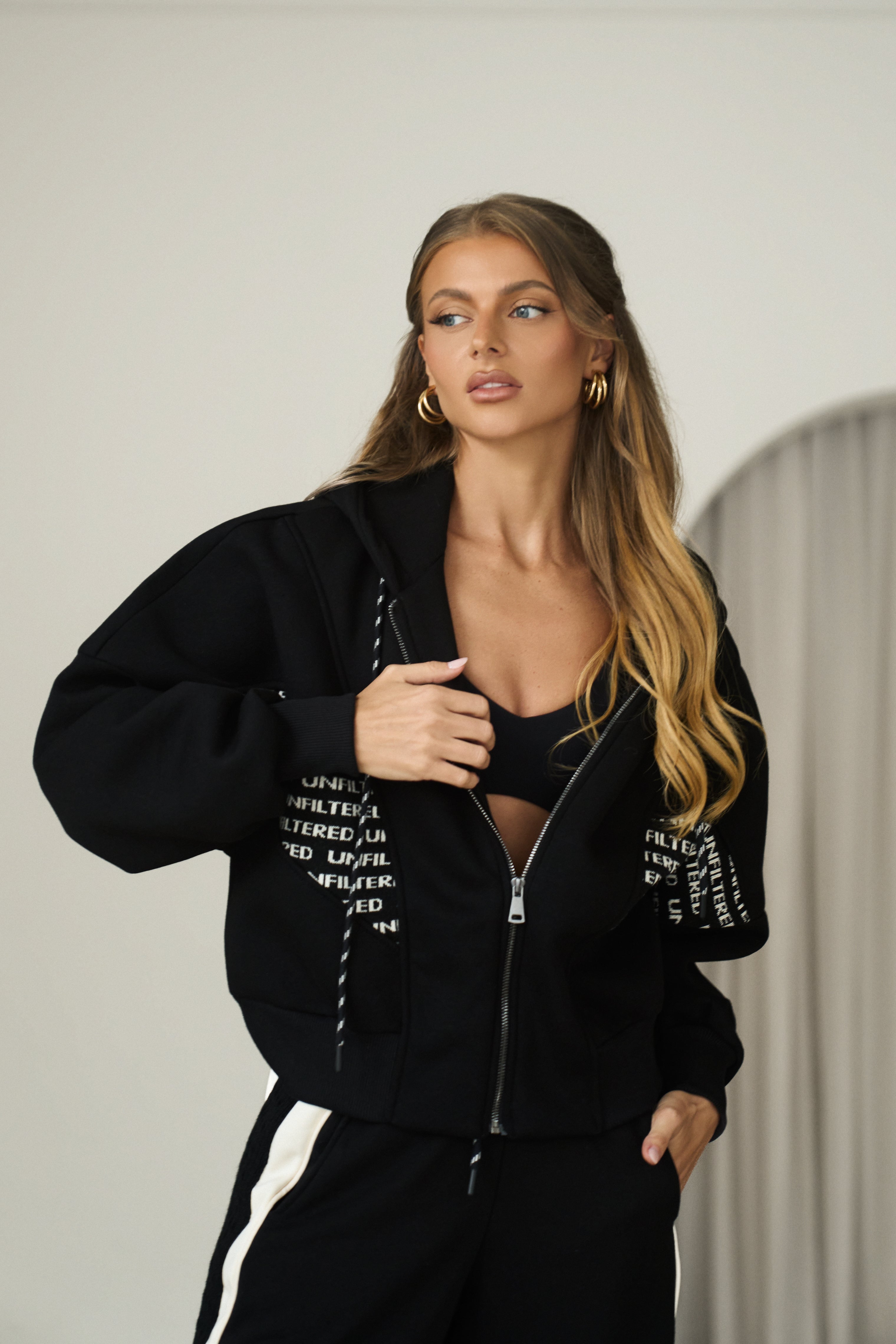 A woman modeling a black zip-up hoodie with white text graphics on the sleeves and chest, paired with black track pants featuring a white side stripe, set against a neutral indoor background.